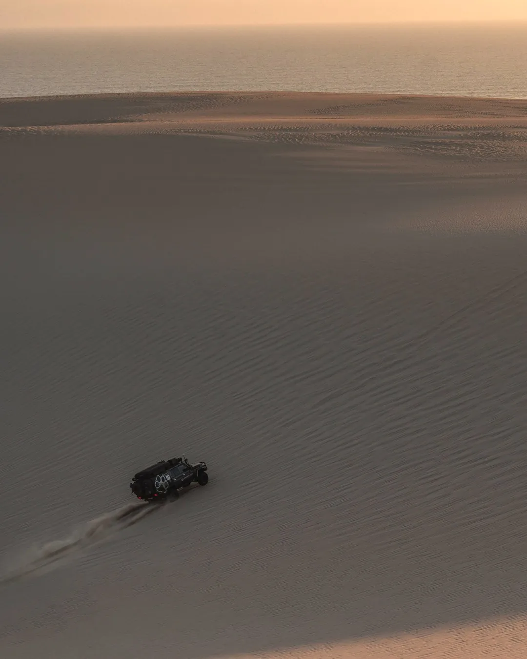 A lone 4×4 climbing a vast sand dune above the coast at dusk